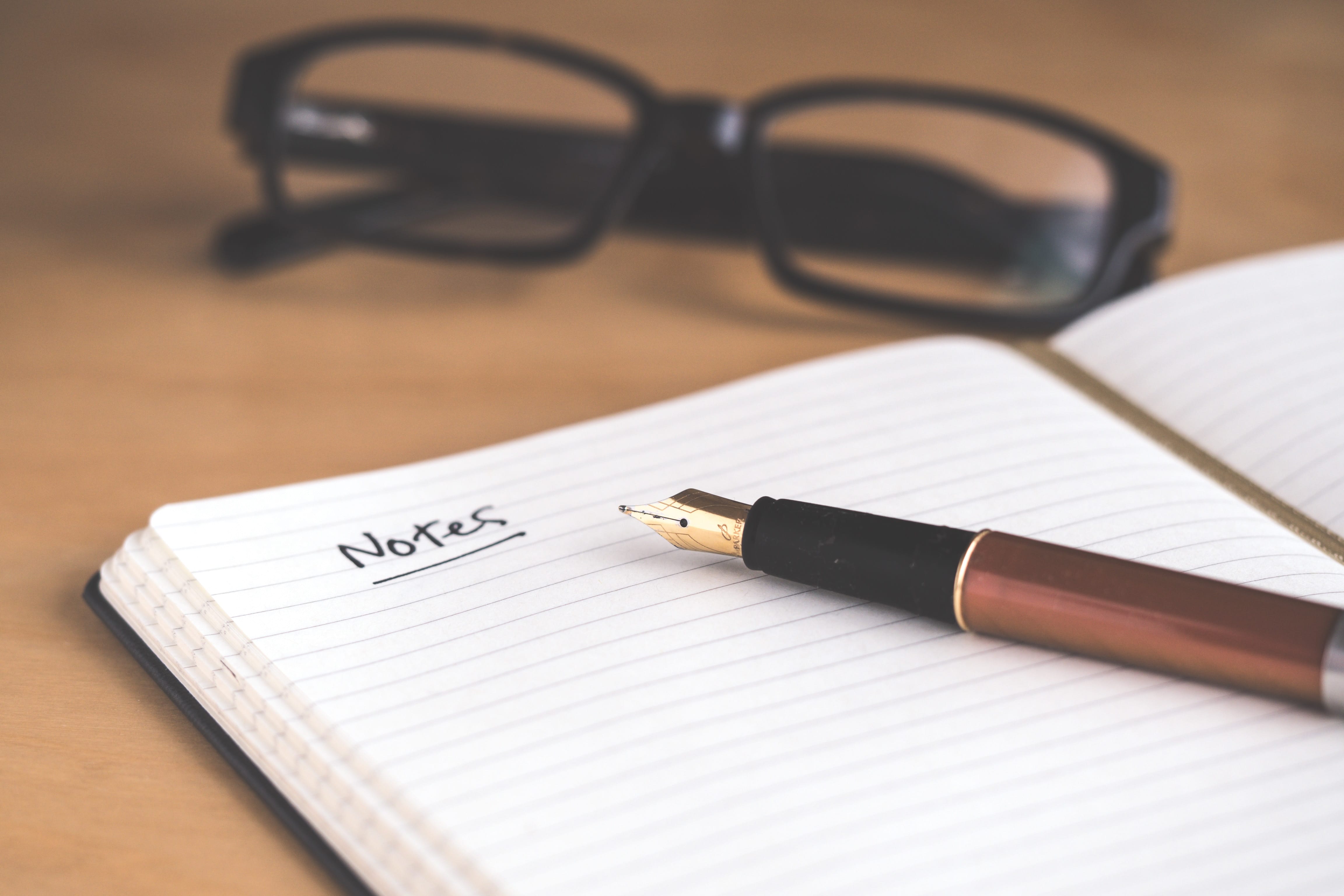Aufgeschlagenes Notizbuch auf einem Tisch mit geöffnetem Füller und Brille im Hintergrund. Auf der Seite steht geschrieben das Wort Notes