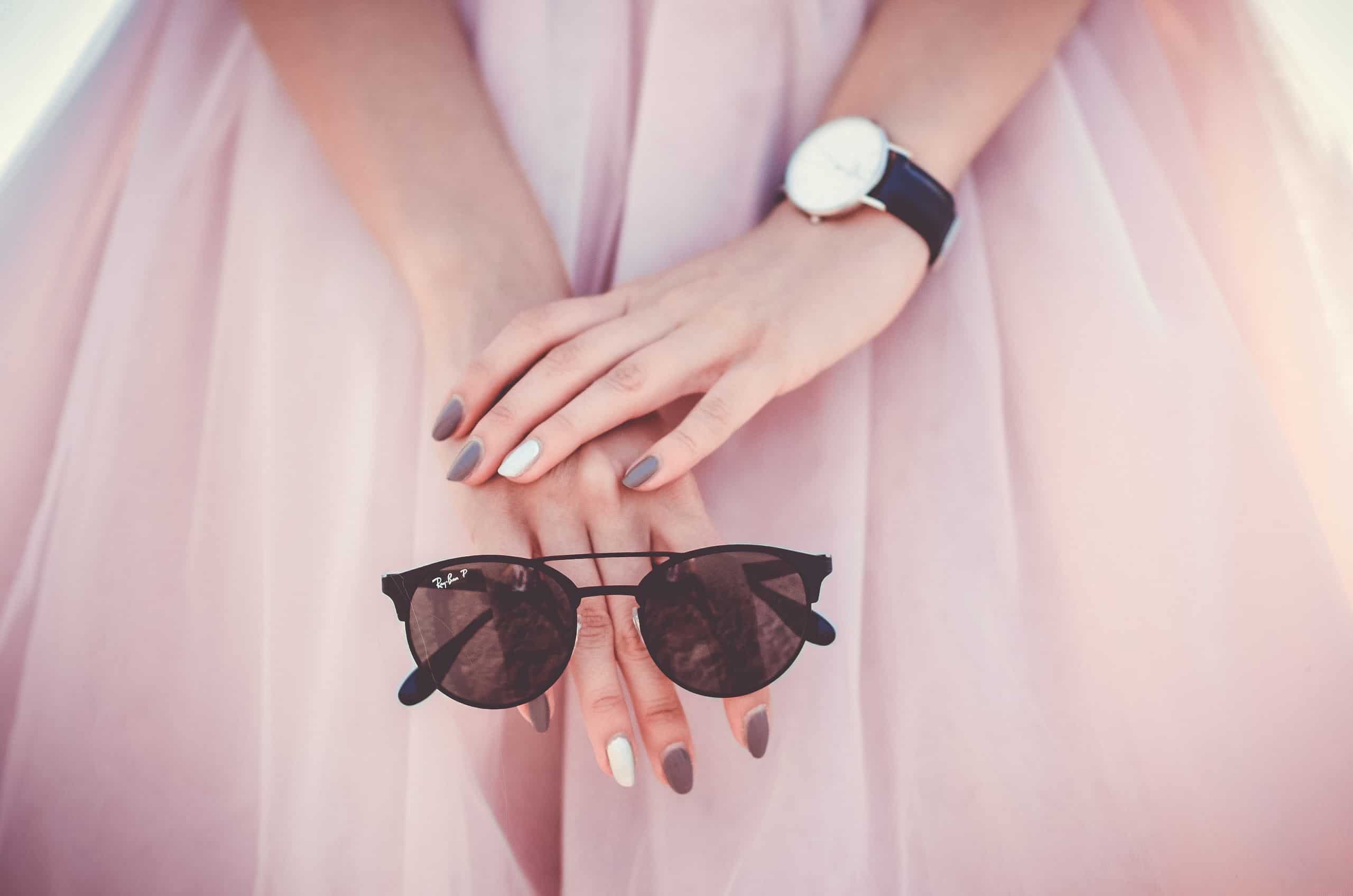 Nahaufnahme von Händen einer Frau, die sie vor einem rosa Tüllrock gefaltet hat. Sie hat eine Sonnenbrille in der Hand und eine Uhr am Handgelenk.
