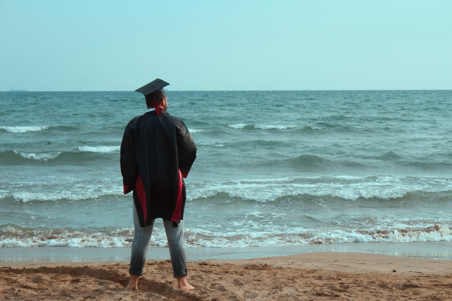 Junger Mann mit Doktorhut auf dem Kopf steht im Sand am offenem Meer