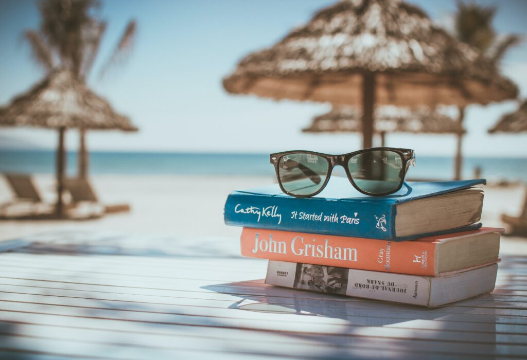 Am Strand ist ein Holztisch mit drei Büchern, die aufeinander gestapelt sind. Darauf liegt eine Sonnenbrille.